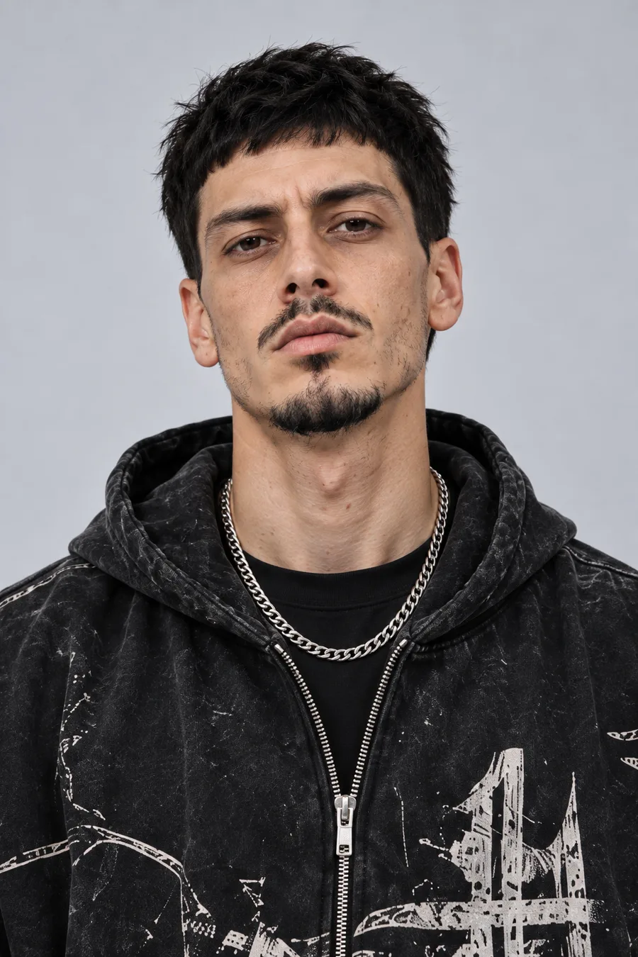Porträtt av MC P: smalt ansikte, mörk mustasch och kort skägg, svart hoodie och silverkedja.